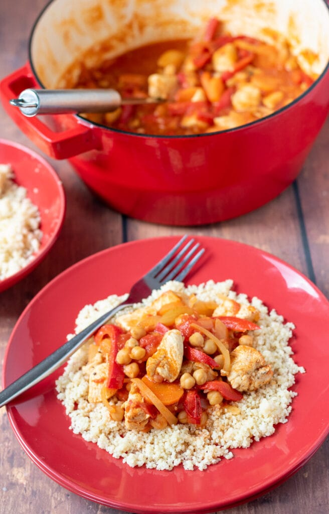 Chicken and chickpea stew served on a bed of couscous with a fork to the left side. Rest of stew in casserole pot at the top with large serving spoon in.