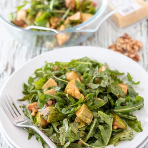 Rocket pear salad with walnuts and balsamic dressing served on a plate with a fork to the side. Rest of salad in a serving bowl, some walnuts and block of parmesan cheese in the background.