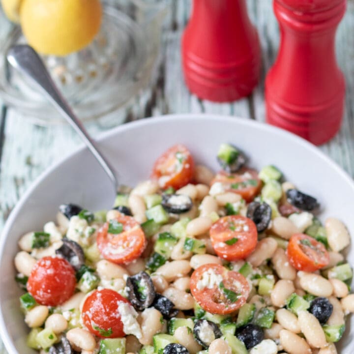 Easy cannellini bean salad served in a round white bowl with a serving spoon in. A lemon and salt and pepper cellars above.