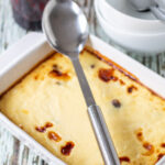 Best baked rice pudding recipe with a serving spoon sitting over the top. Jar of blackcurrant jam and dessert bowls with spoon in at the side.
