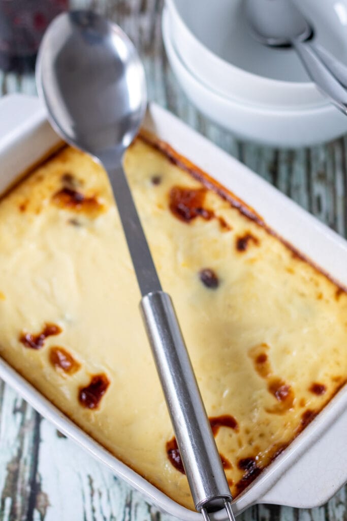 Baked rice pudding removed from oven and sitting with a serving spoon on top. Serving bowls and jar or jam at the top sides.