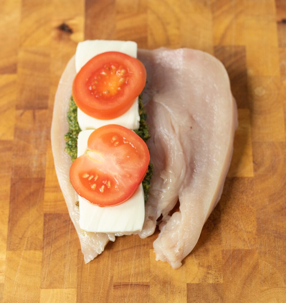 Half a split open chicken breast topped with pesto, mozzarella slices and tomato slices.