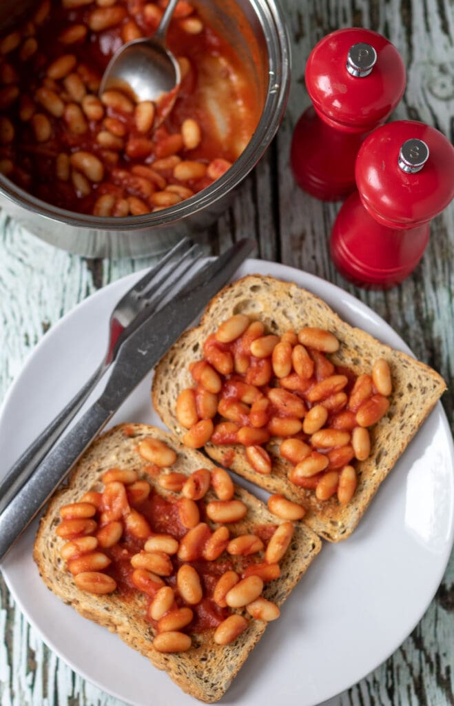 Birds eye view of Easy British Baked Beans on two slices of toast. Knife and fork to the side. Saucepan of homemade beans and salt and pepper cellars above.