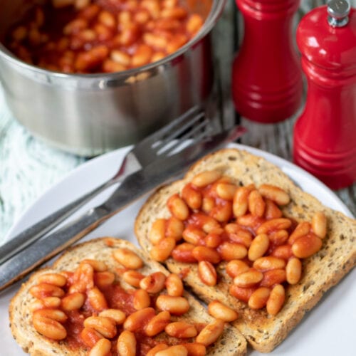 Easy British baked beans served on two slices of wholemeal toast on a white plate. Beans in a saucepan, knife and fork and salt and pepper in the background.
