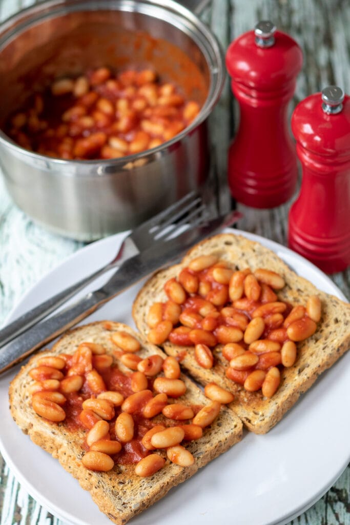 Easy British baked beans served on two slices of wholemeal toast on a white plate. Beans in a saucepan, knife and fork and salt and pepper in the background.