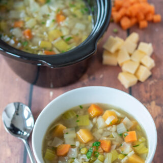 A bowl of easy slow cooker scotch broth with a spoon beside. Rest of the scotch broth recipe in a slow cooker in the background. Diced vegetables scattered around.