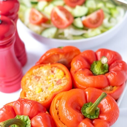 Healthy quinoa stuffed peppers in a white dish. One with the top off. Salt and pepper cellars to the side and a bowl of salad in the background.