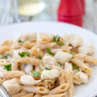 White sauce chicken pasta recipe. Low fat chicken pasta in white sauce on a plate garnished with chopped parsley. A glass of wine and salt cellar in the background.