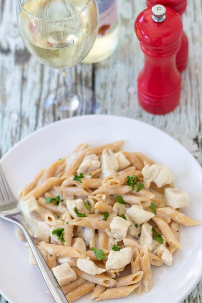 Birds eye view of a plate of low fat chicken pasta in white sauce garnished with parsley. Fork to the side. Glass of wine and salt and pepper cellars above.