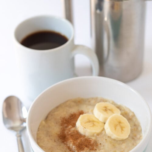 Quick and easy banana porridge served in a bowl garnished with sliced banana and ground cinnamon. Serving spoon to the left side. Mug of coffee and cafetiere in the backbround.