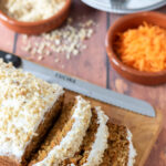Birds eye view of best carrot cake loaf on a bread board with three slices cut away. Plates, grated carrot and chopped nuts arranged as decoration above.