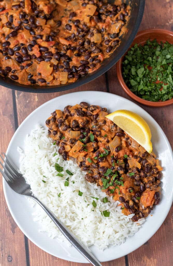 Birds eye view of a plate of black bean curry served with white rice and a fork to the side. Rest of curry in a large pot above with a dish of chopped coriander in between.