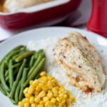 Baked Cajun stuffed chicken breast served on a bed of rice with sweetcorn and green beans as accompaniment on a plate. More chicken breasts in a casserole dish in the background with salt and pepper cellars in between.