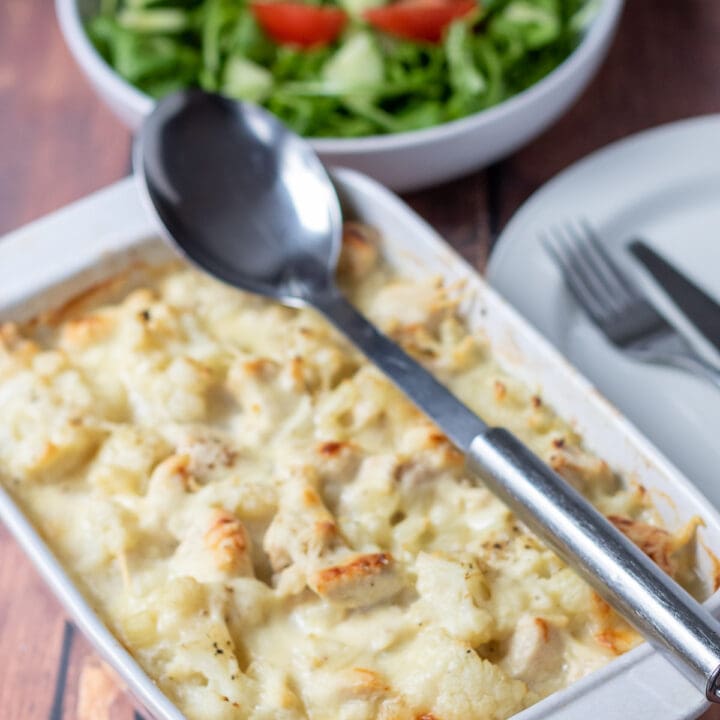Baked chicken and cauliflower cooked in a casserole dish and removed from the oven. Serving spoon on top. Green salad in a bowl at the back with a plate and knife and fork to the side.