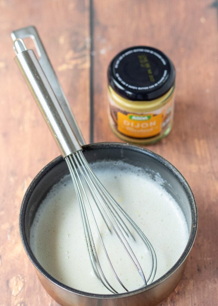 White sauce and a whisk in a small saucepan. Jar of Dijon mustard at the rear.