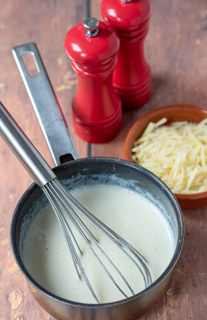Cheese added to white sauce in a saucepan to create cheese sauce. Salt and pepper cellars and dish of grated cheese to the side.