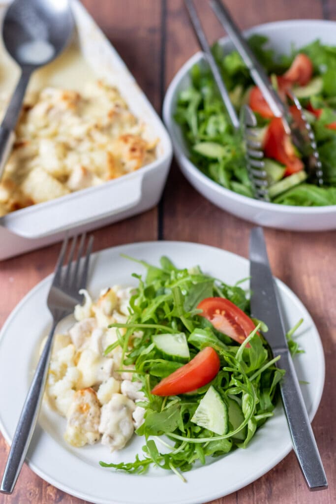 A portion of baked chicken and cauliflower served on a plate with salad. Knife and fork either side of the plate. Rest of the chicken and cauliflower bake in the background alongside a bowl of salad.