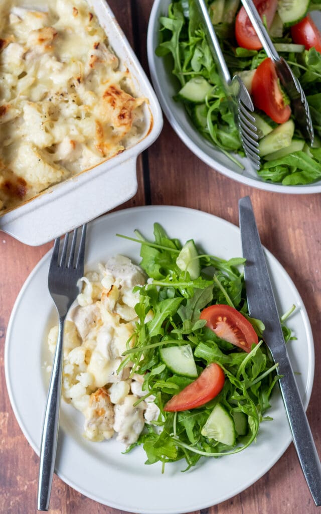 Overhead view of a plate of baked chicken and cauliflower casserole served on a plate with a green salad. Rest of casserole above with salad in a bowl alongside.