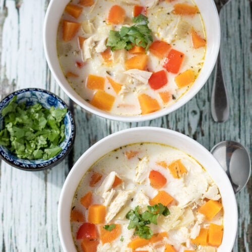 Overhead view of 2 bowls of chicken coconut curry soup garnished with chopped coriander. Dish of chopped coriander to the left side.
