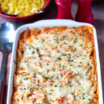 Easy creamy chicken pasta bake in a large red casserole dish with serving spoon to the left. Bowl of sweetcorn and broccoli in the background along with salt and pepper cellars.