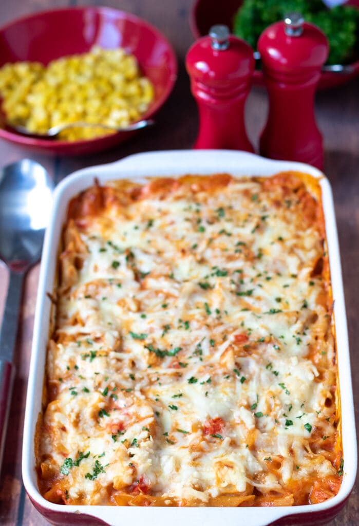 Easy creamy chicken pasta bake in a large red casserole dish with serving spoon to the left. Bowl of sweetcorn and broccoli in the background along with salt and pepper cellars.