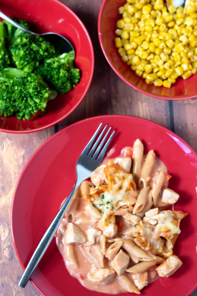 Birds eye view of a plate of creamy chicken pasta bake with a fork to the side. Bowl of broccoli and a bowl of sweetcorn above.
