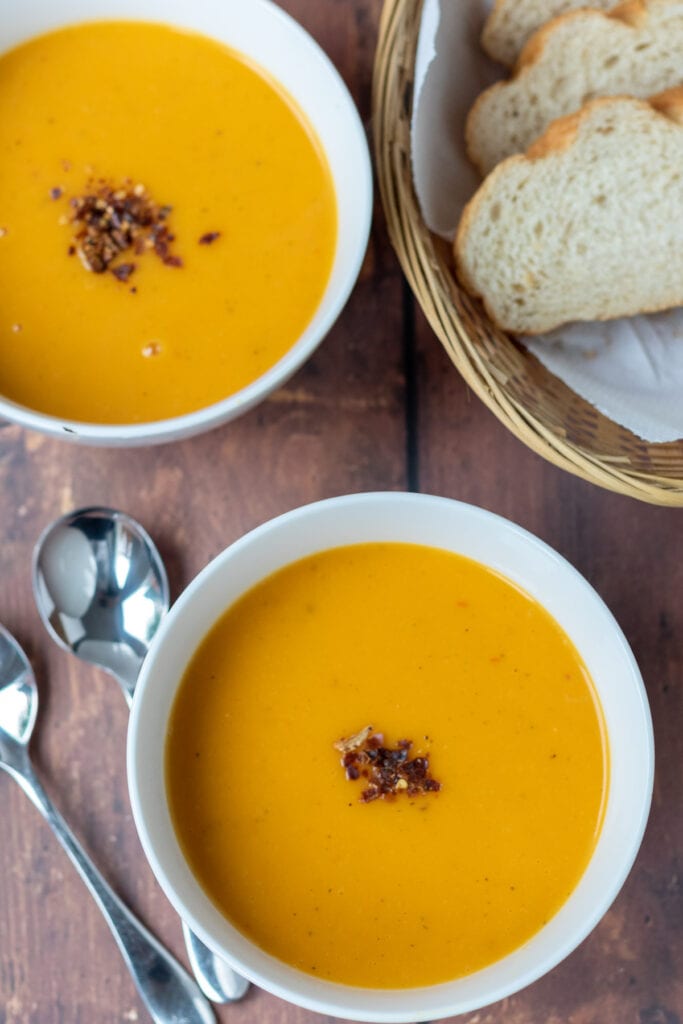 Overhead view of two bowls of roasted spicy butternut squash soup diagonally across from each other. Soup spoons and a basked of slices of bread to each side.