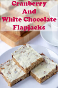 Three cranberry and white chocolate flapjacks on a serving plate. More flapjacks cut on a board in the background. Pin title text overlay at top.