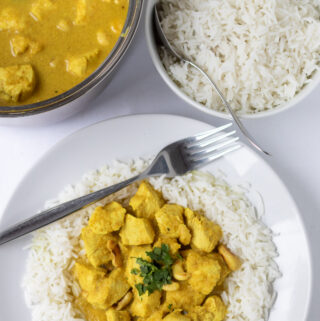 Overhead view of a plate of easy chicken cashew curry served on basmati rice garnished with chopped coriander. Fork to the side. Above a bowl of rice and rest of curry in a saucepan.