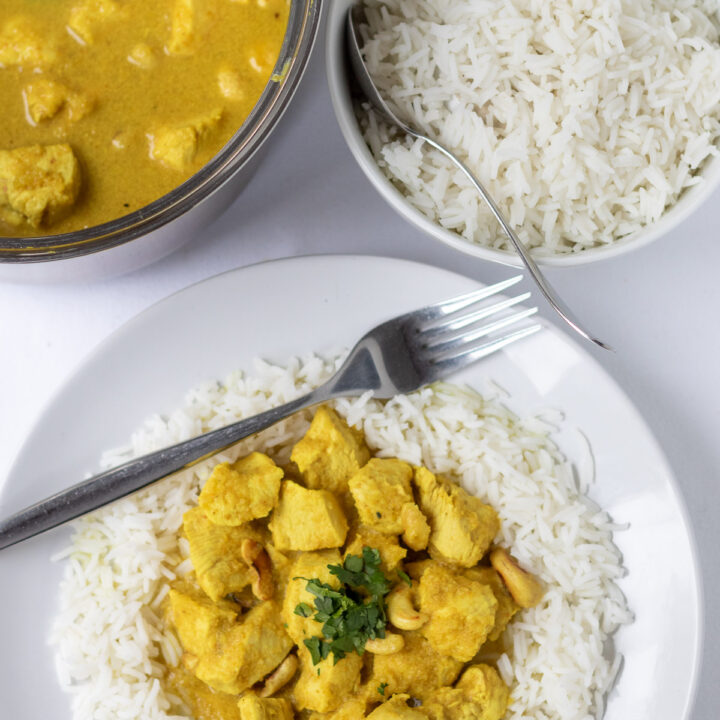 Overhead view of a plate of easy chicken cashew curry served on basmati rice garnished with chopped coriander. Fork to the side. Above a bowl of rice and rest of curry in a saucepan.