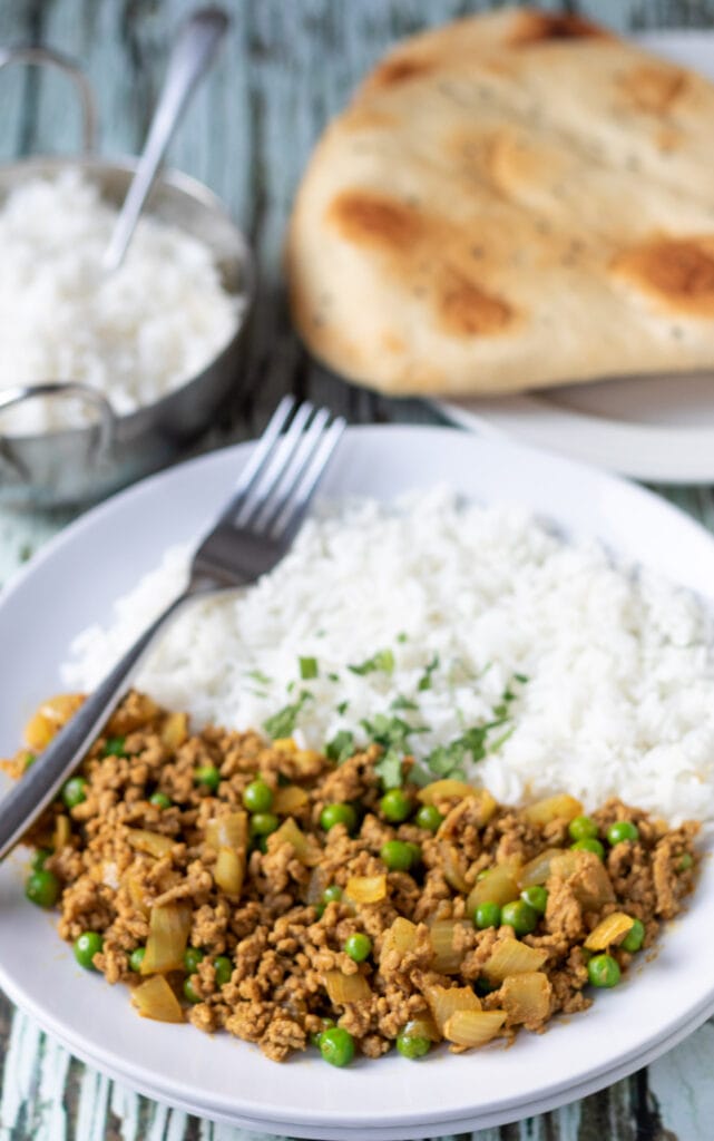 A plate of lamb mince curry served with rice and a fork to the left hand side. A balti dish of rice and a plate of naan breads in the background.