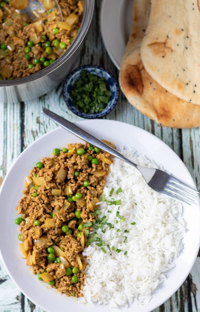 Overhead view of a plate of easy lamb mince curry served with basmati rice and garnished with chopped coriander, fork to the side. Saucepan and plate of naan breads above.