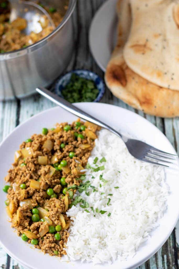 Lamb mince curry served with plain basmati rice and garnished with chopped coriander. Naan breads and rest of curry in pan in the background.
