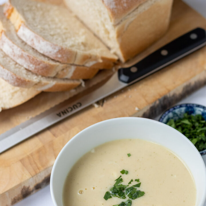 A bowl of low carb roasted cauliflower soup garnished with freshly chopped coriander with a soup spoon beside and loaf of sliced bread in the background.