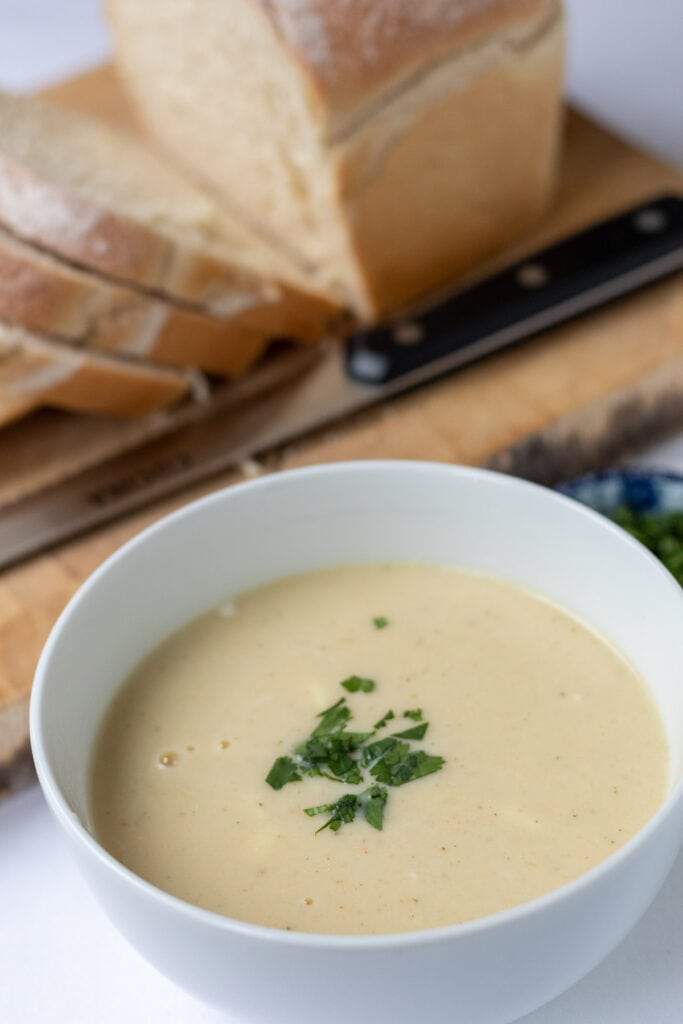 Close up of a bowl of low calorie cauliflower soup garnished with chopped coriander in front of a loaf of sliced bread.