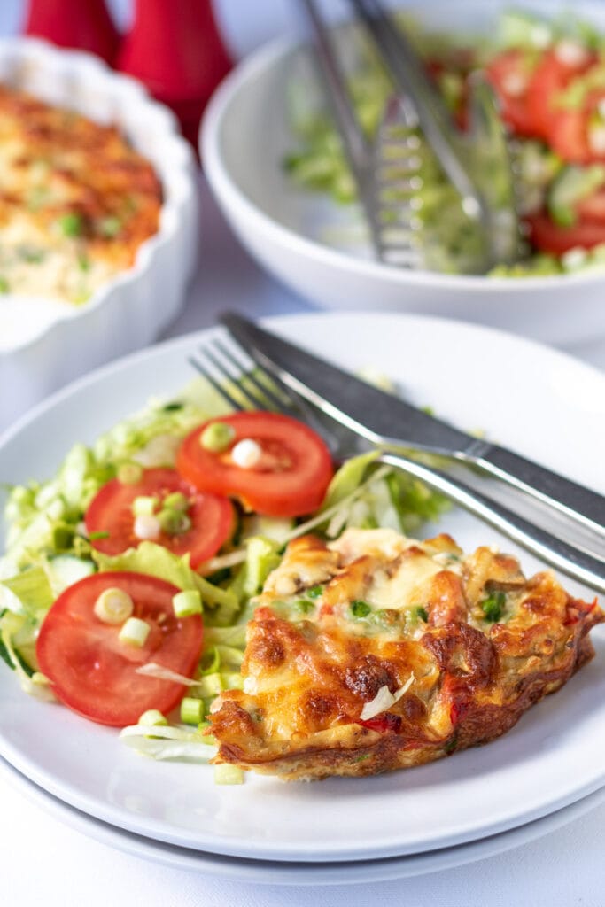 A slice or oven baked vegetable frittata served on a plate with a salad. Knife and fork to the side. Salad bowl and frittata dish in the background.