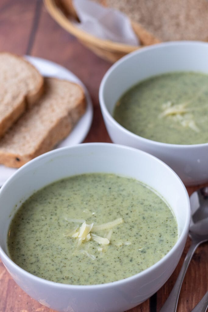 Two bowls of healthy creamy kale soup one in front of the other. A plate with two slices of bread on to the left and soup spoons to the right.