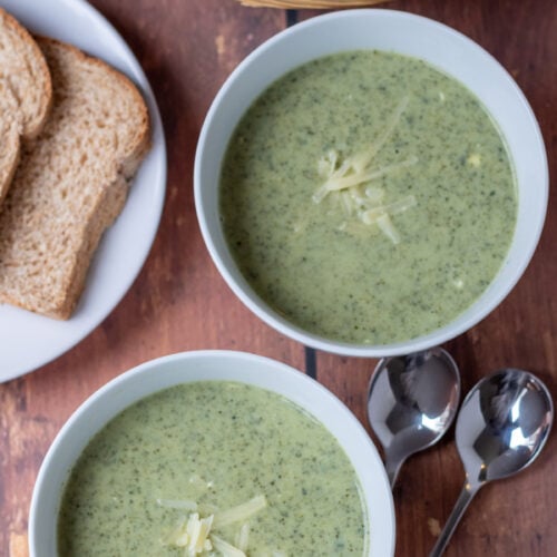Overhead view of two bowls of healthy creamy kale soup garnished with grated cheese. Slices of bread in a bread baked and on a plate and two soup spoons at the side.
