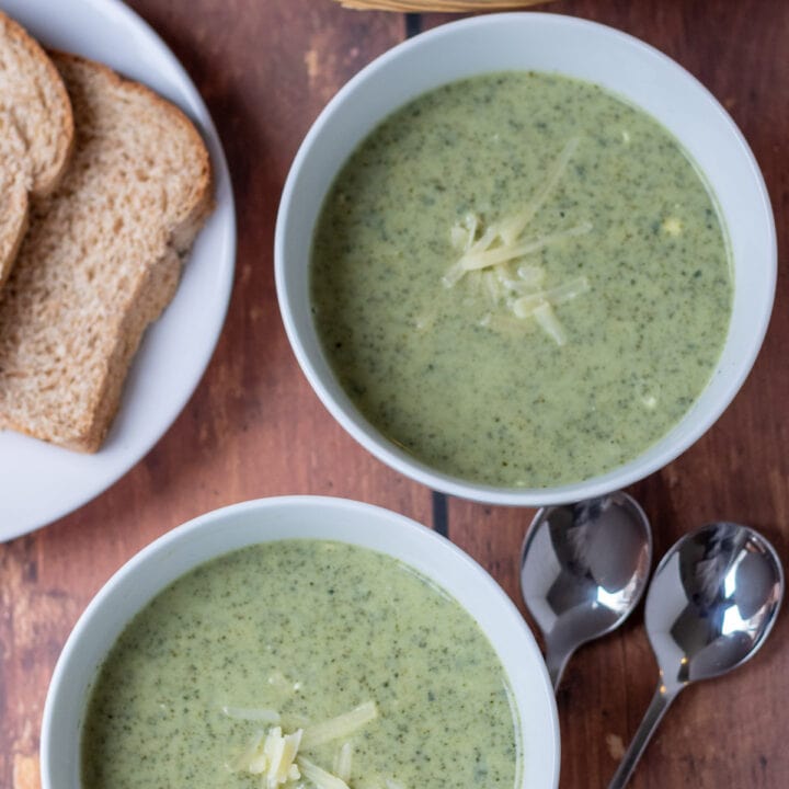 Overhead view of two bowls of healthy creamy kale soup garnished with grated cheese. Slices of bread in a bread baked and on a plate and two soup spoons at the side.