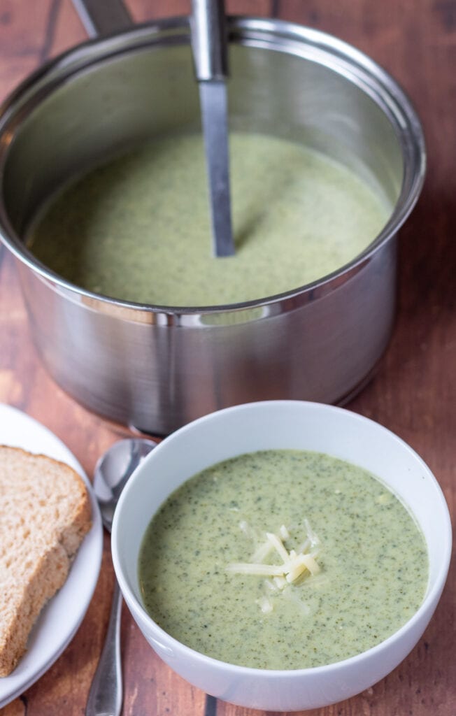 A bowl of healthy creamy kale soup topped with grated cheddar cheese. Rest of soup in a saucepan with a ladle in behind. Slices of bread on a plate to the left with a soup spoon in between.