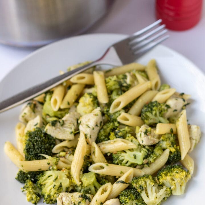 A plate of pesto chicken broccoli pasta with a fork. A saucepan and salt and pepper cellars behind.