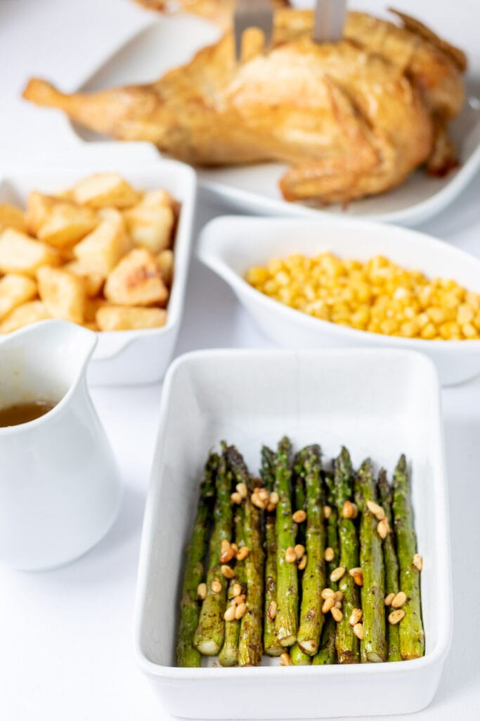 Baked balsamic asparagus served in a square baking dish. Other side dishes of roast potatoes and sweetcorn in the background with a roast chicken at the back.