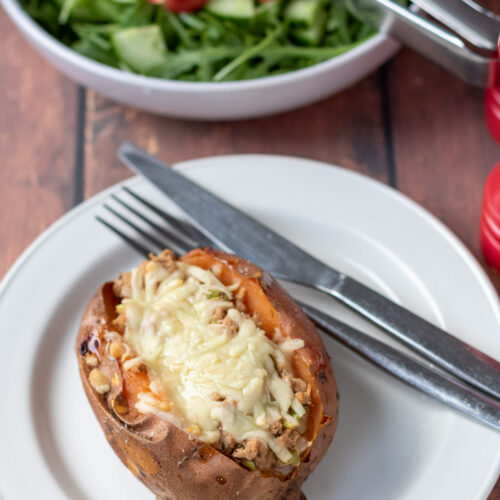 A healthy baked potato with tuna and cheese filling on a plate with a knife and fork to the side. Bowl of salad with sald tongs in at the top.