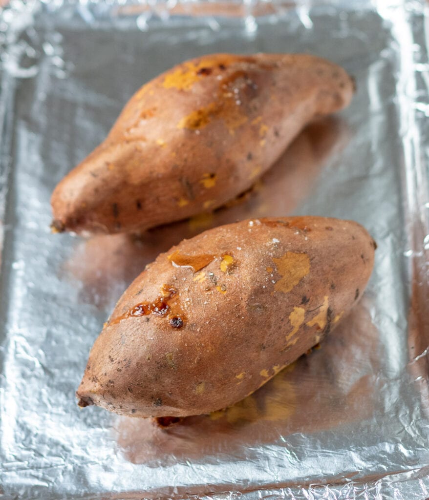 Two sweet potatoes baked in the oven, removed and on an aluminium foil covered baking tray.