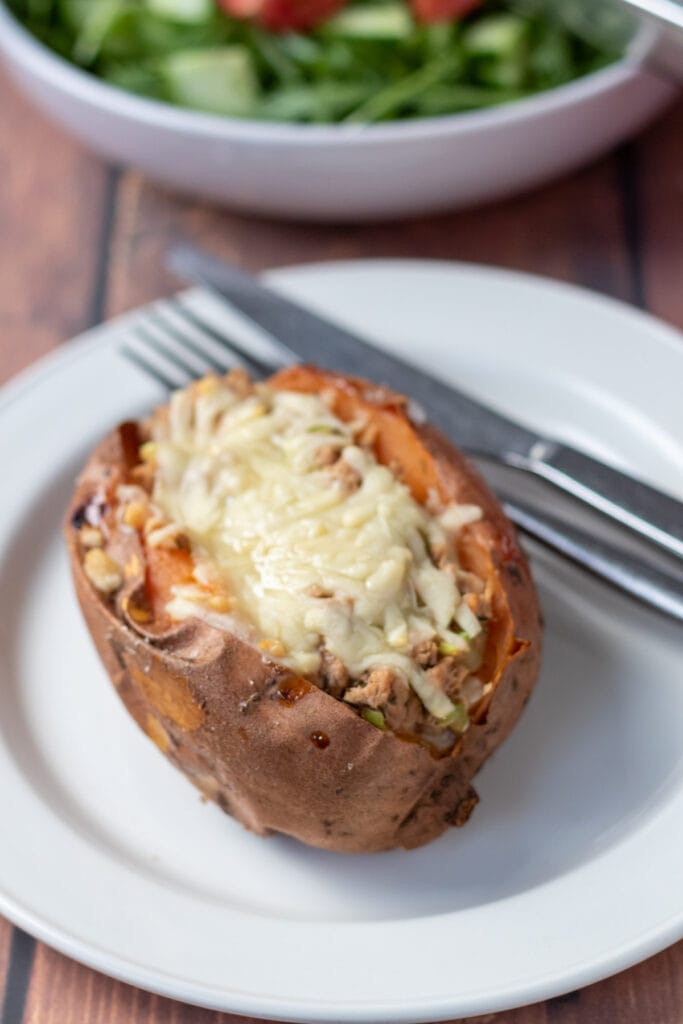 Healthy baked potato with tuna and cheese on a serving plate. Knife and fork to the right hand side. Dish of green salad in the background.