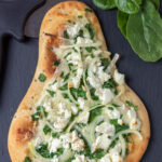 Overhead view of a cooked spinach and feta naan pizza with a pizza cutter and some spinach leaves alongside.