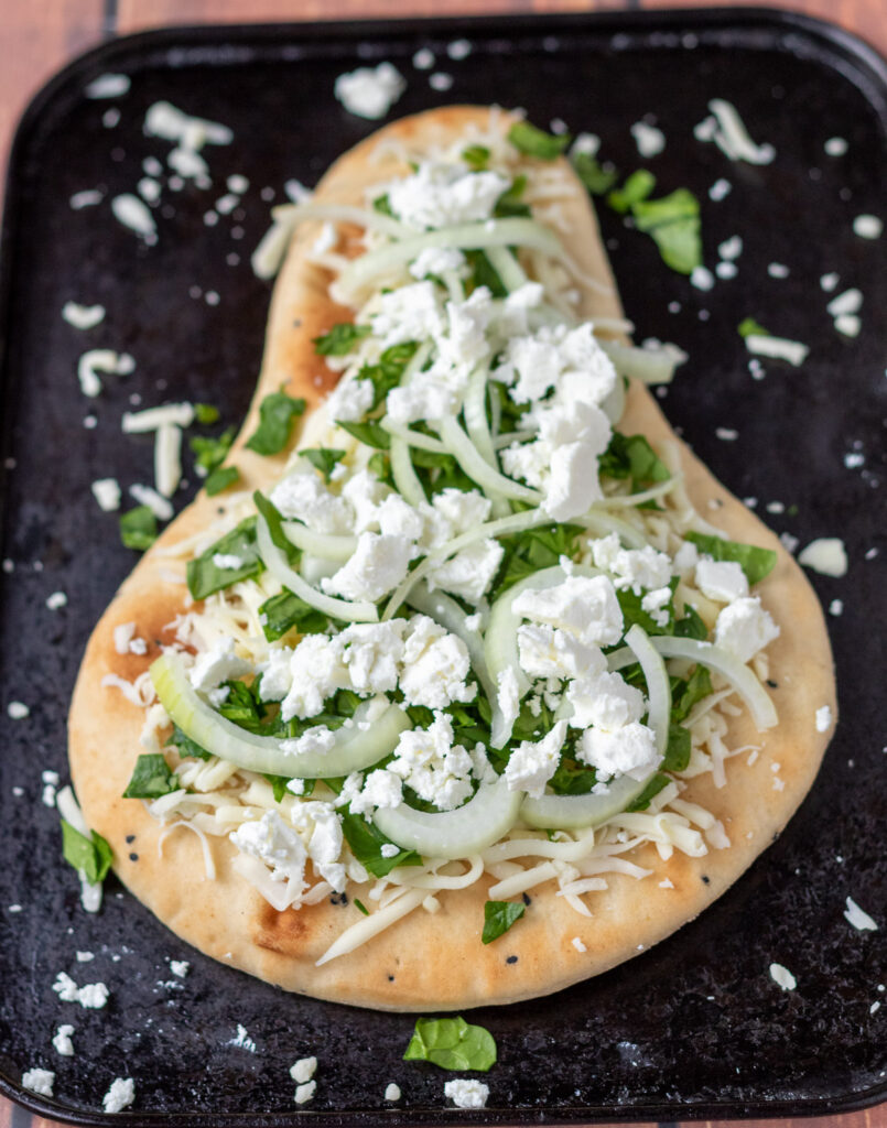 Naan bread on a baking tray sprinkled with garlic, mozzarella, spinach, onion and feta cheese.