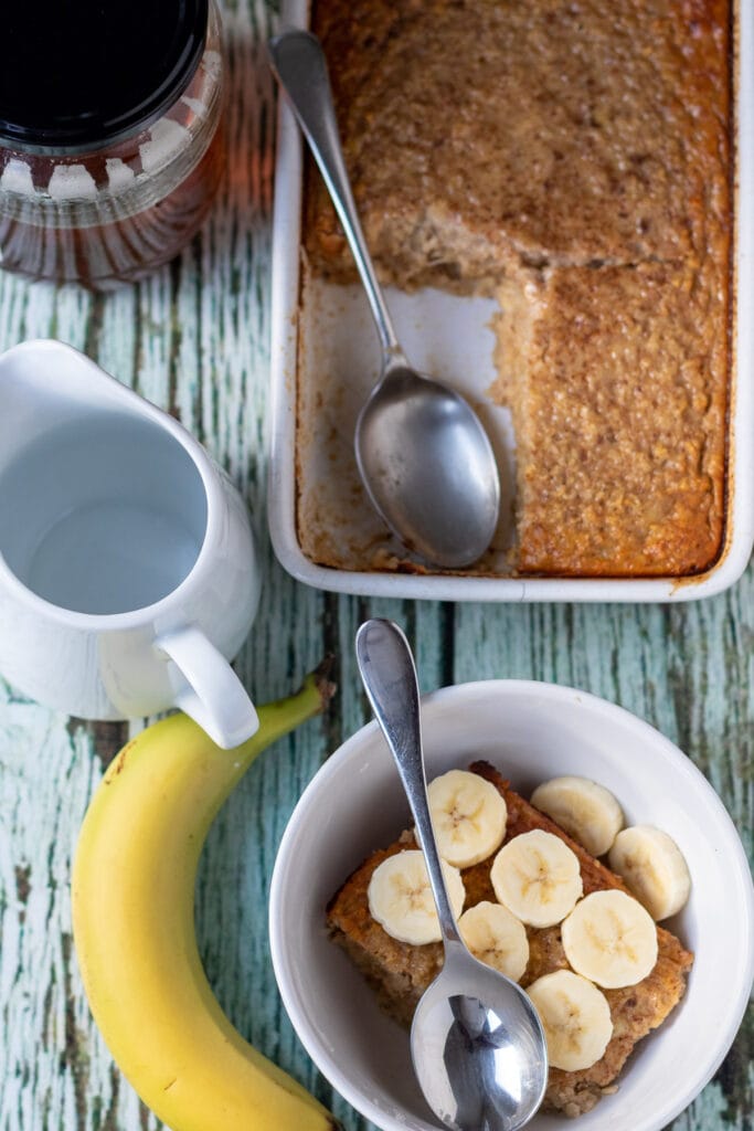 Overhead view of a bowl of banana baked oats topped with slices bananas. A banana to the side and rest of recipe still in serving dish at top.