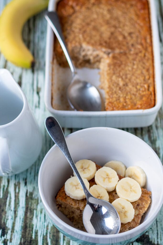 A bowl of served baked banana oatmeal decorated with sliced bananas. Rest of the recipe in casserole dish above.