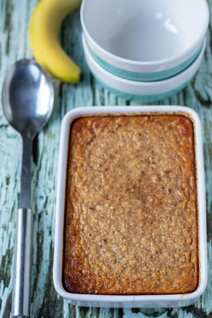 Baked banana oatmeal just removed from the oven and still in cooking dish. Serving spoon to the left and serving bowls above.